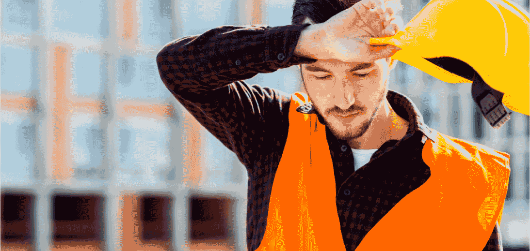 Trabajador en obra afectado por calor extremo, con chaleco de seguridad y casco en mano, representando riesgo de golpe de calor laboral.
